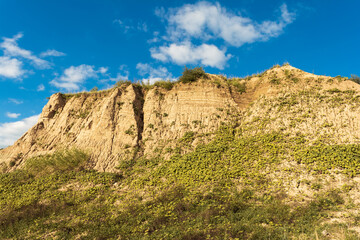 Sandy cliff covered with green vegetation under a bright blue sky with scattered clouds, showcasing a natural landscape with rugged terrain and plant growth.