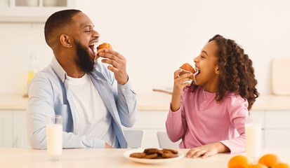 It's Breakfast Time. Portrait of cheerful black little girl and man eating sweet muffin cake cookies and looking at each other, dad and daughter sitting at table in kitchen, enjoying meal with milk