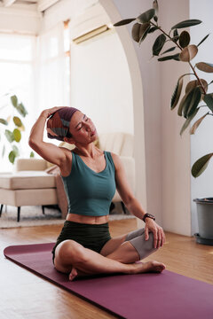 Woman practices yoga, stretching neck on mat indoors.