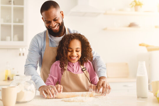 Home Bakery Concept. Happy african american man and his child daughter rolling up dough with pin together, having fun, daddy and daughter making cookies, cake, bread or preparing pizza, copy space - Powered by Adobe