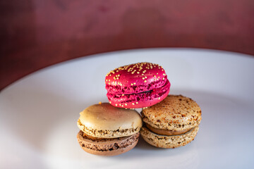 Colorful macarons on white floral plate against warm textured background. Red, beige, and chocolate tones with visible fillings. Cozy and inviting dessert still life.