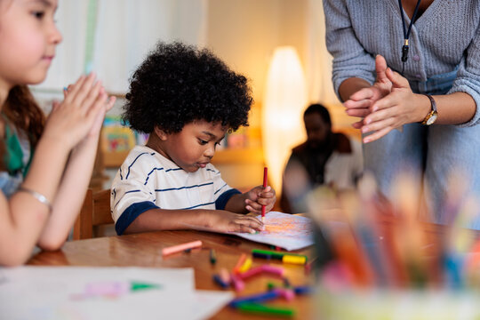 Children drawing with crayons, teacher clapping in classroom.