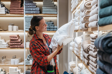 Female shopper in a cozy plaid shirt examines a fluffy white towel in a well-organized retail store, surrounded by neatly stacked linens and soft textures, showcasing home goods