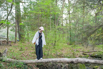 Fototapeta premium A woman is walking on a log in a forest. She is wearing a white shirt and a hat