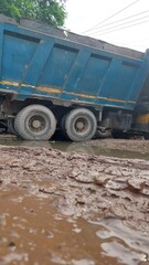 Fototapeta premium Curated image of blue dump truck stuck in a muddy puddle on a dirt road after heavy rain.