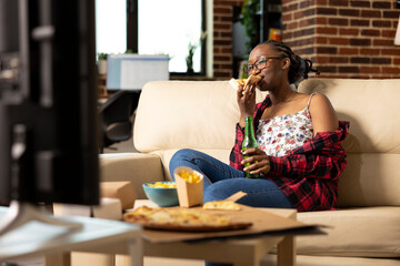 Portrait of relaxed woman enjoying slice of pizza while watching TV in brick wall apartment. Black female individual sitting comfortably on sofa, having quiet movie night with favorite fast food.