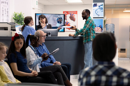 Hospital staff assists black man at reception desk while female doctor consults elderly male patient in clinic lobby. Receptionist multitasks with phone call and paperwork in busy waiting area.