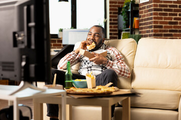 African american male individual relaxing at home during weekend, eating juicy burger and watching TV. Young black man having casual afternoon in apartment with fast food and entertainment on screen.