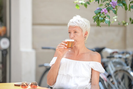 A woman in a white shirt is drinking a beer while sitting at a table. The scene is casual and relaxed, with the woman enjoying her drink and the outdoor setting - Powered by Adobe