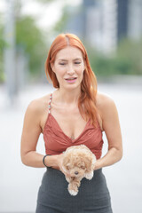 A woman with red hair is holding a small dog. The woman is wearing a red top and black skirt