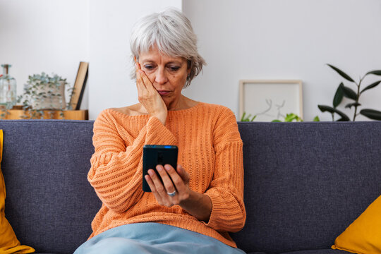 Worried senior woman reading bad news on smart phone message sitting on sofa at home.