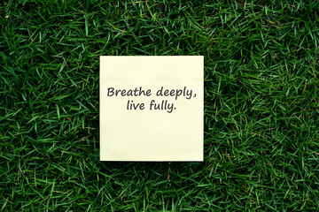 Yellow sticky note with inspirational quote "Breathe deeply, live fully." placed on fresh green grass background. Motivation, mindfulness, and positive lifestyle concept.
