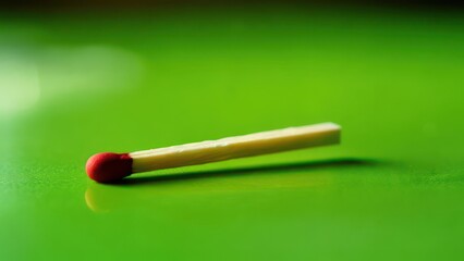 Close-up of a single matchstick lying on a vibrant green reflective surface, slightly out of focus in the background. The match head is red, and the wooden stick is pale yellow