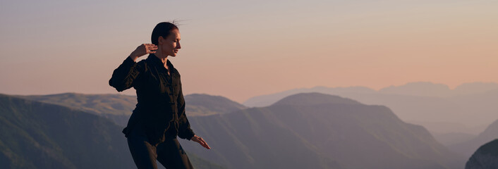 Woman practicing Tai Chi in traditional black outfit on mountain at sunrise, arms outstretched in...