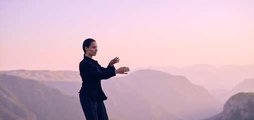 Woman practicing Tai Chi in traditional black outfit on mountain at sunrise, arms outstretched in...