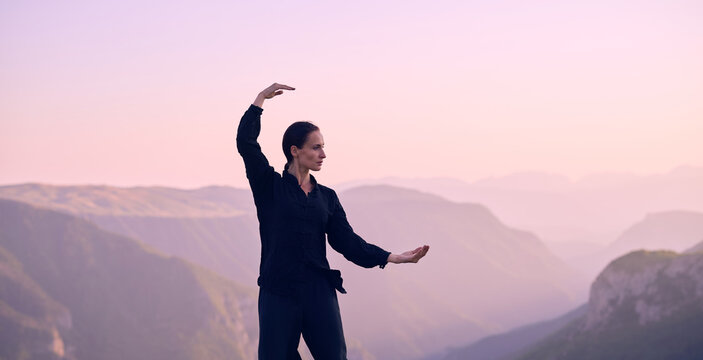 Woman practicing Tai Chi in traditional black outfit on mountain at sunrise, arms outstretched in meditation pose focusing on balance, energy, and mindfulness in nature.