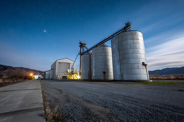 Obraz premium Grain silos and industrial building under starry sky with mountains in background at dusk