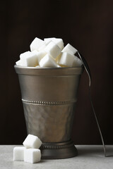 A pewter cup filled with sugar cubes witha spoon leaning on the cup and three cubes on the table.