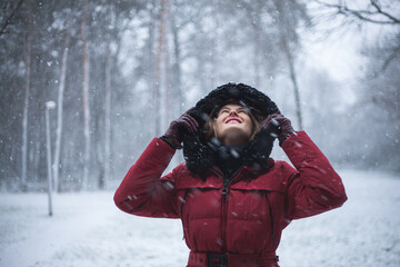 Portrait of beautiful young woman in red winter hooded jacket, while looking and and cheerful watching snowflakes falling all around. Fashion model in winter scene forest, snowing, Christmas, season.