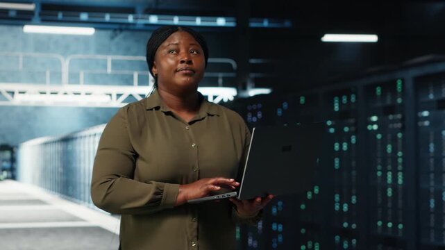 Admin walks between server farm rows, using laptop to upgrade rigs for increase in clients workload capacity. Data center worker configures hardware with notebook, improving processing speed, camera B