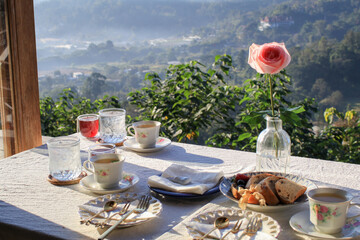 Breakfast table with homemade Whole grain bread, hot herbal tea and flower in vase with fog cover mountain. Morning nature view.