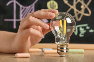 Female hand with light bulb and chalk on wooden table against chalkboard with science drawings, closeup. Inventor concept