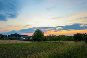 ​A rural landscape at sunset, where multicolored clouds stretch across the sky, illuminating grassy fields and trees. The view of the village in the distance highlights the peace and idyll of country 