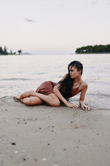 A young woman lies on wet sand by the shore in a brown swimsuit, gazing toward the horizon as gentle waves surround her in a tranquil beach moment.