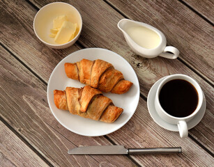 Two fresh croissants black coffee butter and cream on a wooden table.