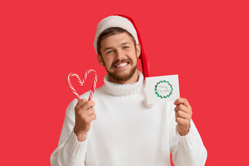 Happy young man in Santa hat with Christmas greeting card and candy canes on red background