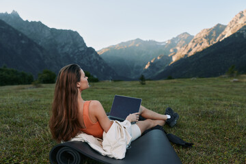 A woman sits outdoors on a yoga mat with a laptop in a scenic mountain meadow. She is relaxed yet focused, blending work with nature in a tranquil, inspiring setting.