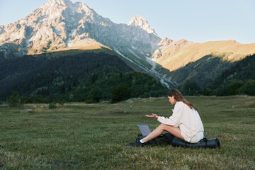 Naklejka premium A lone traveler sits on a grassy field with a laptop, immersed in work as dramatic mountains rise behind, creating a serene outdoor workspace.