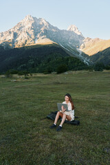 Fototapeta premium A focused student sits on a grassy field with a laptop, surrounded by rugged mountains. She studies outdoors, enjoying fresh air and sunlight, creating a calm and productive learning atmosphere.