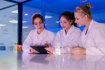 A team of three young female scientists smiles while working together in a futuristic, blue-lit lab. They are looking at a tablet, sharing ideas and discussing their research.