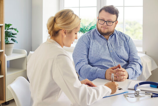 Doctor and patient during obesity consultation support. The physician writes notes as the fat overweight man listens during medical exam in clinic. Collaborative medical weight management