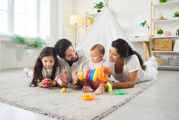 Happy father, mother, small girl and her sister lying together on floor, playing with colorful pyramid, talking to each other, posing in childrens room, spending leisure time in family circle