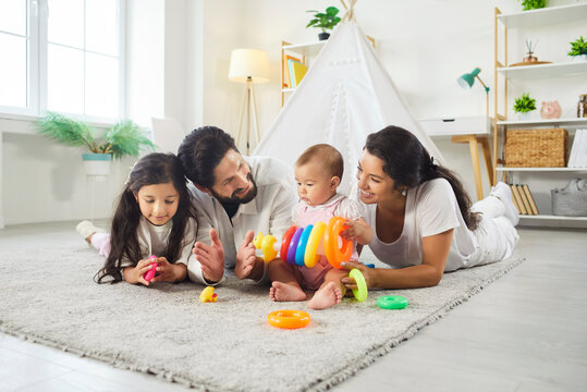 Happy father, mother, small girl and her sister lying together on floor, playing with colorful pyramid, talking to each other, posing in childrens room, spending leisure time in family circle - Powered by Adobe