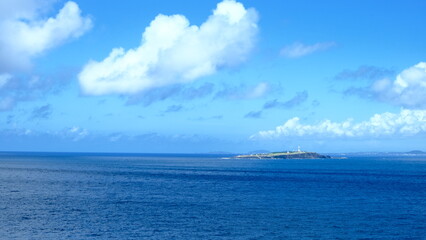 A serene seascape featuring an island with a lighthouse in the distance, under a sky filled with fluffy clouds. 