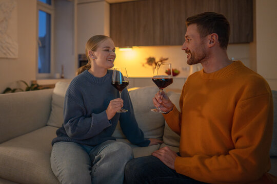 Laughing couple drinking wine on sofa looking at each other lovingly in intimate light. Romantic evening, emotional connection, love relationship, man and woman bonding at home, date on weekends