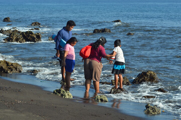 Four family members remain near the shoreline, calmly watching waves break and foam against the rocks while holding supplies in bright daylight.