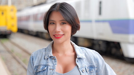 Young woman in denim jacket standing on a train platform, looking directly at the camera with blurred trains in the background