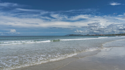 Beautiful seascape. The long waves of the ocean roll towards the shore, foam, and spread over the sandy beach. Silhouettes of mountains, hotel buildings on the horizon. Blue sky, clouds. Malaysia. 