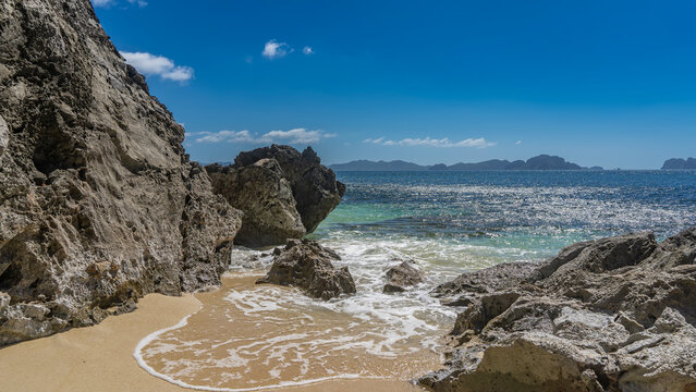 A secluded beach on a tropical island. Picturesque coastal cliffs. The waves of the turquoise ocean spread over the sand. Delicate foam. Silhouettes of mountains on the horizon. Blue sky, clouds.  