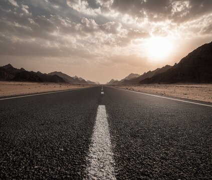 A straight asphalt road stretches through a desert landscape with mountains in the distance under a cloudy sunset sky