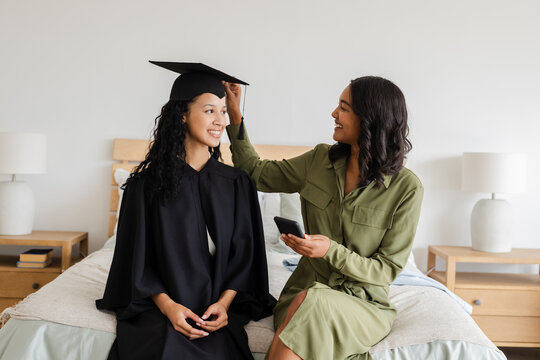 Proud mother adjusting daughter's graduation cap while sitting on bed at home