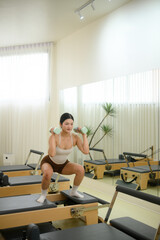 Fototapeta premium Vertical image of an Asian woman training with Pilates squat and weights for powerful strength on the reformer, elegant posture, and mindful movement