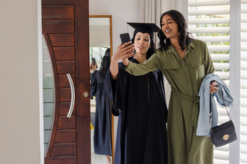 Mother and daughter taking selfie at home, celebrating graduation in cap and gown