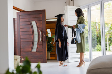 At home, African American mother congratulating daughter in cap and gown on graduation day