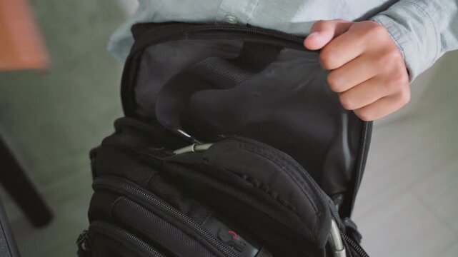 boy in casual shirt holding open black school bag while preparing for school, organizing supplies with focused hands, ready to insert books and materials, indoors near table in clean modern space