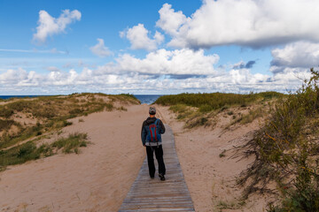 Male hiker walking on wooden boardwalk through sand dunes towards the beach under blue sky with cumulus clouds, peaceful coastal landscape, perfect for travel, nature and outdoor adventure projects